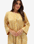 A woman modeling a long-sleeved mustard yellow and beige paisley printed tunic dress with a buttoned front and pleated bodice, set against a plain light gray background.