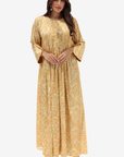 A woman modeling a long, loose-fitting golden-yellow dress with a subtle tonal floral pattern, three-quarter bell sleeves, and a round neckline, set against a plain light gray background.