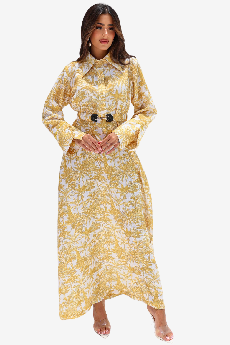 A woman modeling a long-sleeved yellow and white tropical print maxi dress with a matching belt featuring two black buttons, set against a plain light gray background.