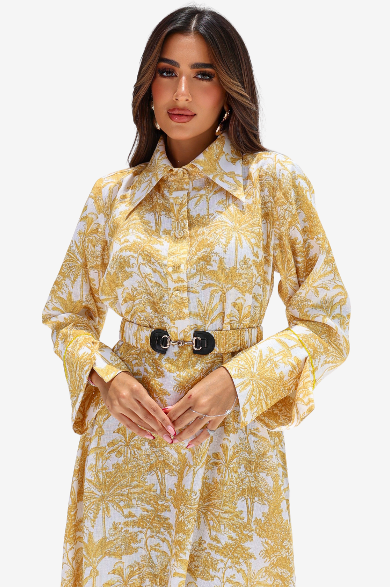 A woman modeling a long-sleeve collared shirt dress with a yellow and white palm tree pattern, featuring a matching belt with large black oval buckles, set against a plain light gray background.