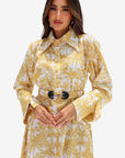 A woman modeling a long-sleeve collared shirt dress with a yellow and white palm tree pattern, featuring a matching belt with large black oval buckles, set against a plain light gray background.