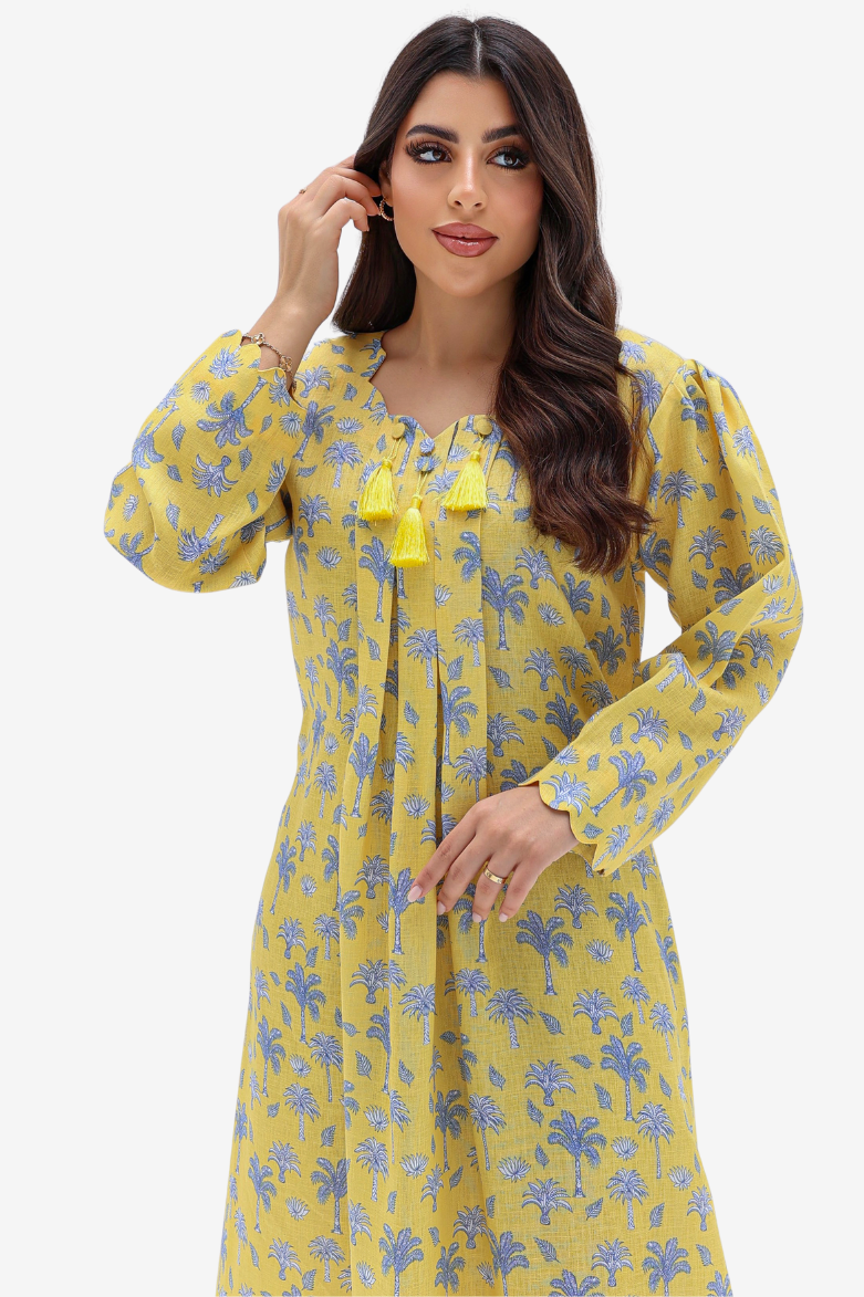 A woman modeling a yellow long-sleeve tunic dress with a blue floral print and yellow tassel tie details at the neckline, set against a plain light gray background.