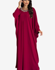 A woman modeling a long, flowing magenta maxi dress with wide draped sleeves and a decorative silver-toned collar, set against a plain light gray background.