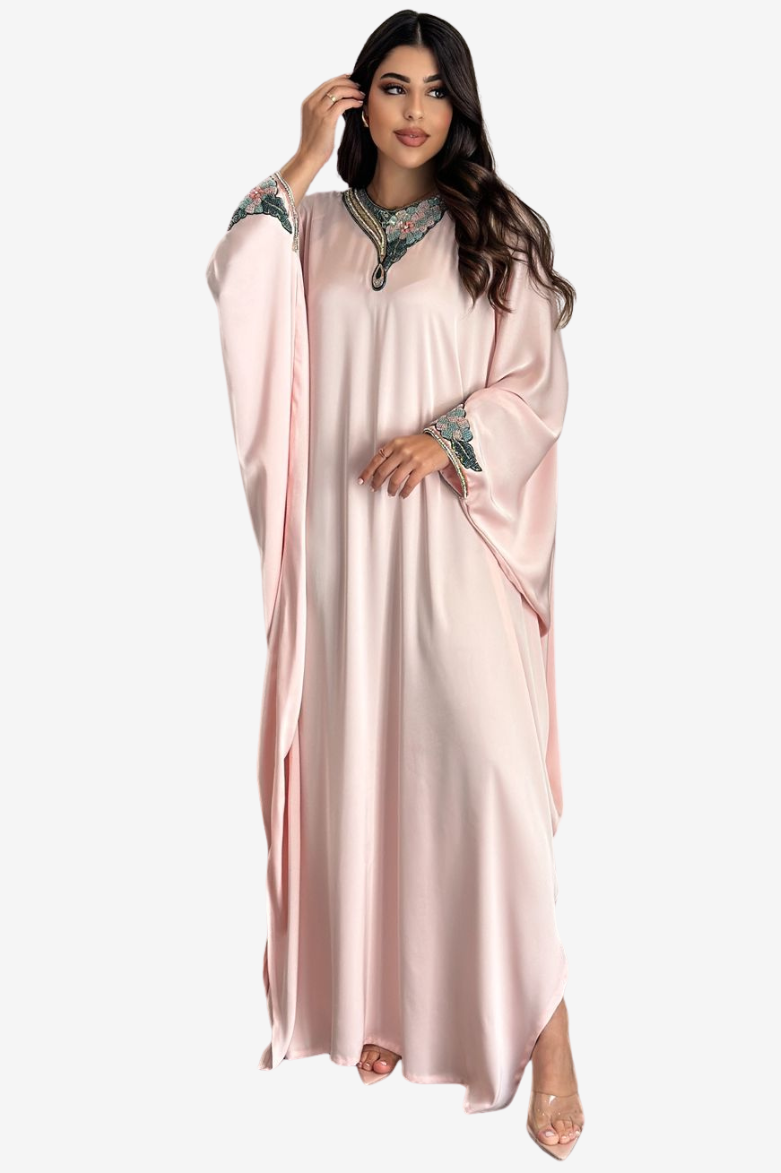 A woman modeling a long, flowing light pink kaftan dress with wide sleeves and ornate embroidered detailing around the neckline and cuffs, set against a plain light gray background.