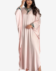 A woman modeling a long, flowing light pink kaftan dress with wide sleeves and ornate embroidered detailing around the neckline and cuffs, set against a plain light gray background.