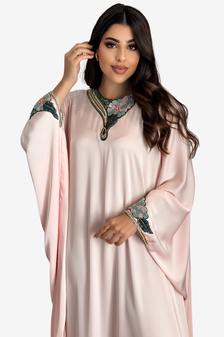 A woman modeling a light pink kaftan dress with embroidered floral details on the collar and cuffs, set against a plain light gray background.