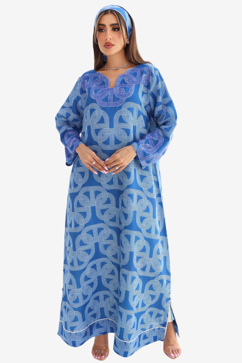 A woman modeling a long blue kaftan dress with a white geometric pattern, paired with a matching blue headband, against a plain light gray background.