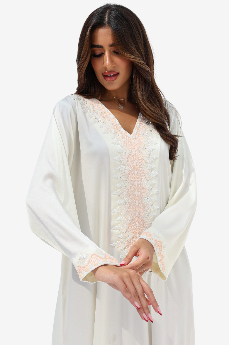 A woman wearing a long-sleeved white kaftan-style tunic with delicate pale pink floral lace embroidery along the V-neckline and cuffs, set against a plain light gray studio background.