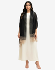 A woman modeling a black sheer open-front cardigan with beige lace trim over a cream maxi dress and nude high heels, against a light gray studio background.
