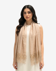 A woman modeling a light beige linen shawl with delicate floral lace trim at the ends, worn over a white sleeveless dress against a plain light gray background.