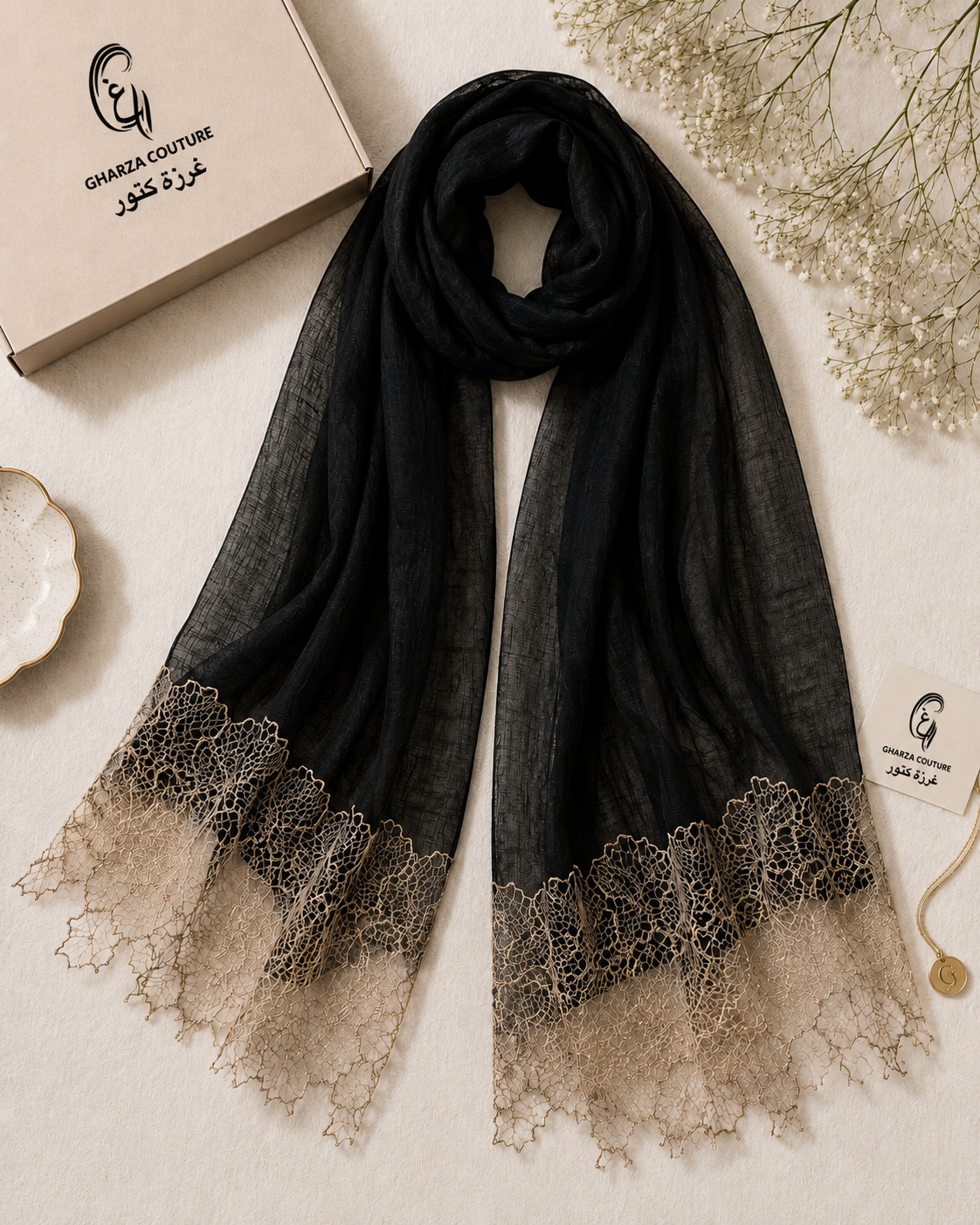 A black sheer scarf with a beige lace hem, styled on a textured beige surface next to its branded box and decorative dried flowers. The brand name 'Gharez Couture' is visible on the packaging and tag.