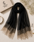 A black sheer scarf with a beige lace hem, styled on a textured beige surface next to its branded box and decorative dried flowers. The brand name 'Gharez Couture' is visible on the packaging and tag.
