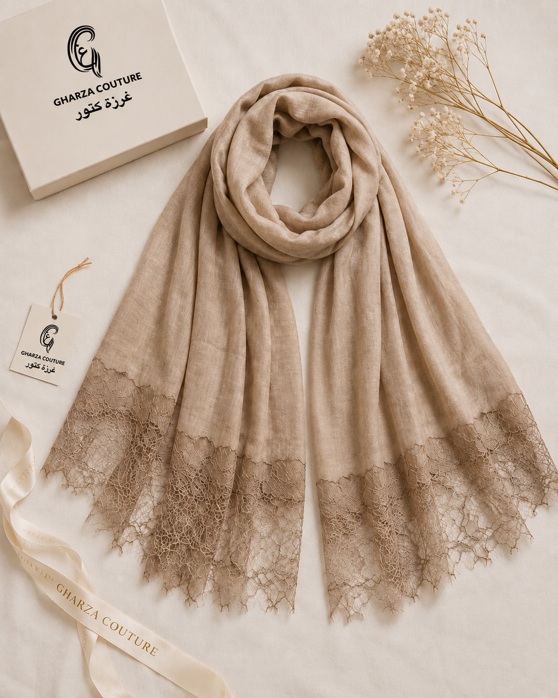 A beige scarf with a delicate lace hem, elegantly draped on a light cream fabric background. A cream-colored box with the brand name 'GHARZA COUTURE' in both English and Arabic is visible in the top left, along with a matching ribbon and dried floral stems.