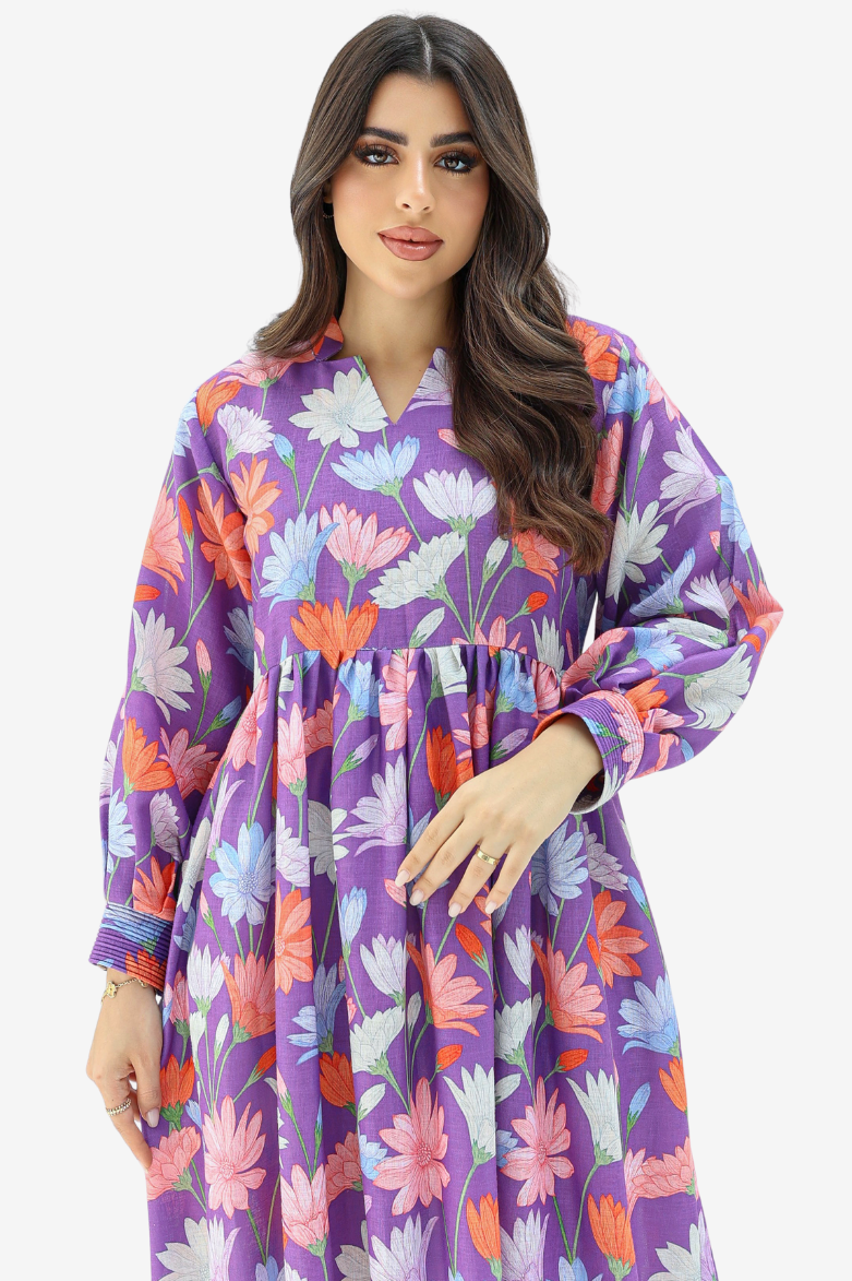 A woman wearing a purple long-sleeve kaftan dress with a colorful floral print in pink, orange, and white, set against a plain light gray background.
