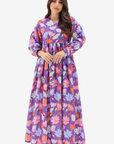 A woman modeling a long-sleeved maxi dress with a purple base and a colorful floral print in pink, orange, and light blue, set against a plain light gray background.