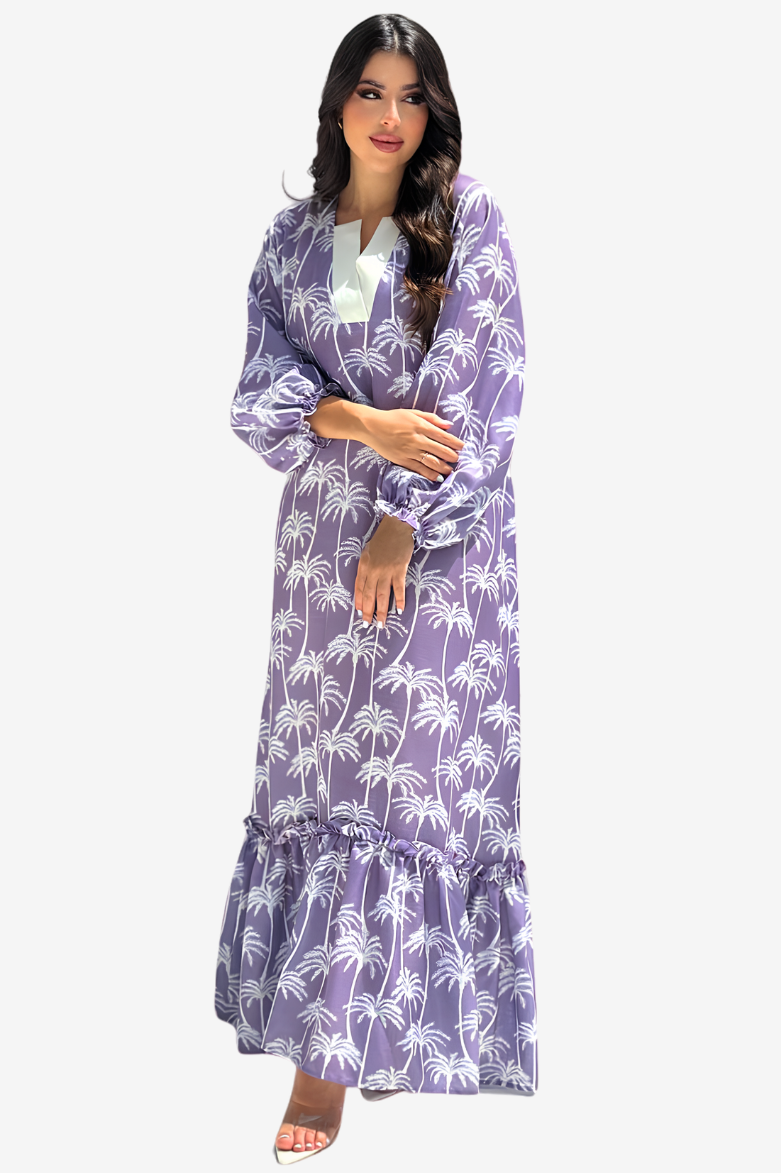 A woman modeling a long-sleeved maxi dress with a purple and white palm tree print, featuring a tiered ruffle hem and a white inner collar, set against a plain light gray background.