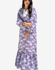 A woman modeling a long-sleeved maxi dress with a purple and white palm tree print, featuring a tiered ruffle hem and a white inner collar, set against a plain light gray background.