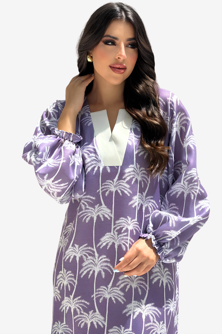 A woman modeling a purple and white palm tree patterned long-sleeve tunic dress with a white V-neckline, set against a plain light gray background.