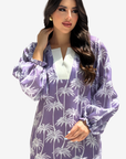 A woman modeling a purple and white palm tree patterned long-sleeve tunic dress with a white V-neckline, set against a plain light gray background.
