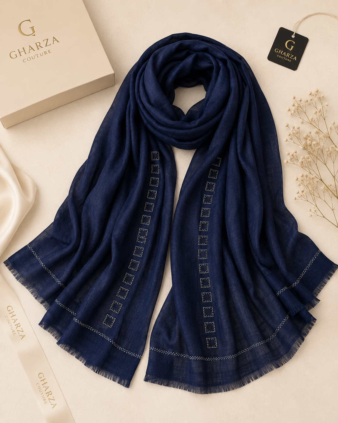 A navy blue Gharza Couture scarf with white geometric embroidered patterns and fringed edges, styled in a flat-lay arrangement with its branded packaging box, ribbon, and hangtag on a light beige background.