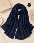 A navy blue Gharza Couture scarf with white geometric embroidered patterns and fringed edges, styled in a flat-lay arrangement with its branded packaging box, ribbon, and hangtag on a light beige background.