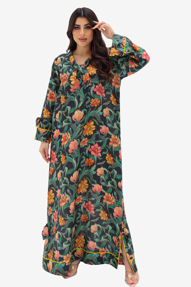 A woman modeling a long-sleeved maxi dress with a vibrant floral print on a dark green background. The dress features orange, pink, and yellow flowers with green leaves, and has a split hem with yellow piping.