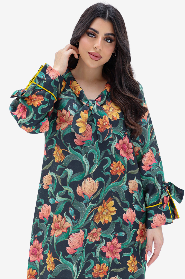 A woman wearing a long-sleeve floral print tunic dress with green leaves, pink and orange flowers, and yellow trim on the cuffs, set against a plain light gray background.