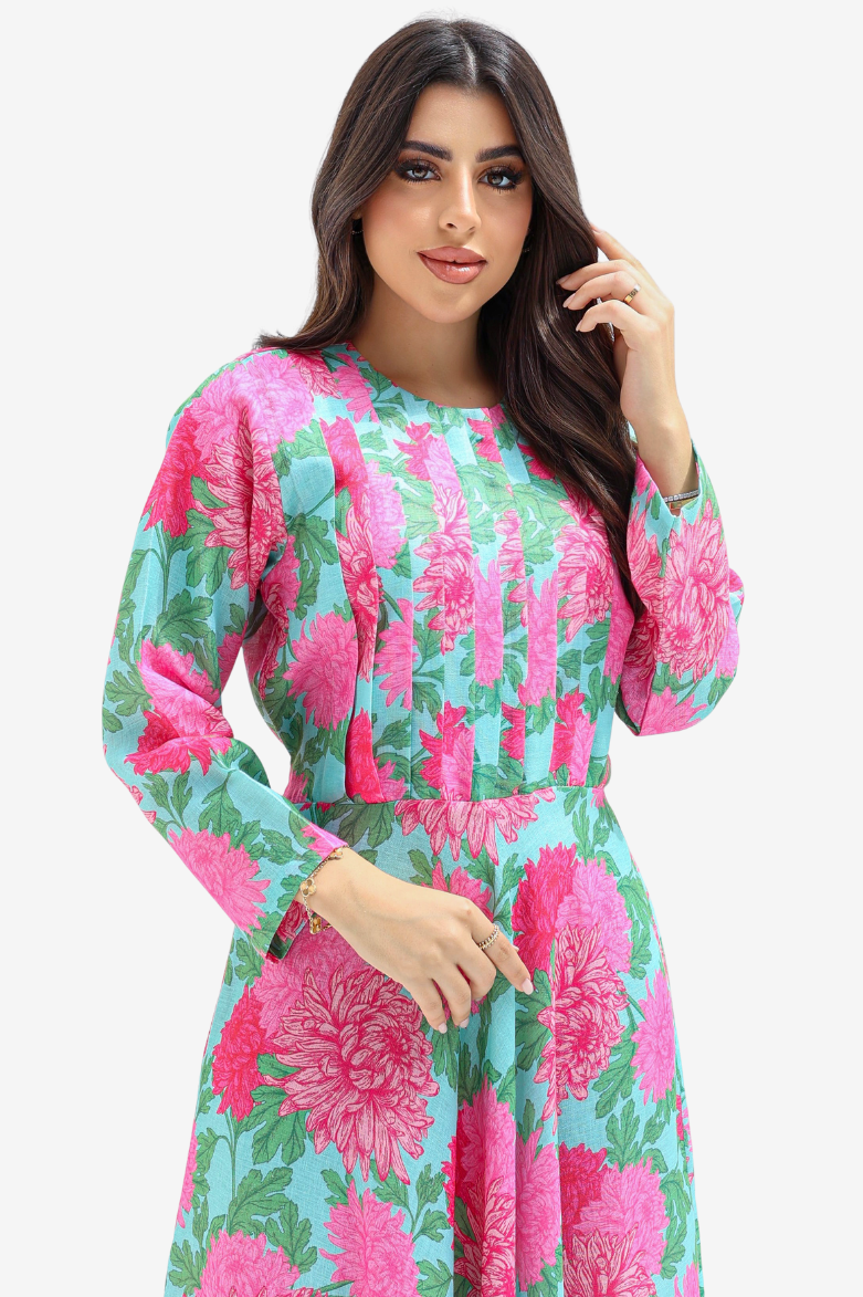 A woman modeling a long-sleeve dress with a vibrant pink and green floral print on a light blue background, set against a plain light gray studio backdrop.