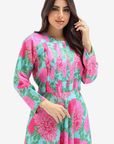 A woman modeling a long-sleeve dress with a vibrant pink and green floral print on a light blue background, set against a plain light gray studio backdrop.