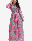 A woman modeling a long-sleeved maxi dress with a vibrant pink and green floral print on a light blue background, set against a plain light gray studio backdrop.