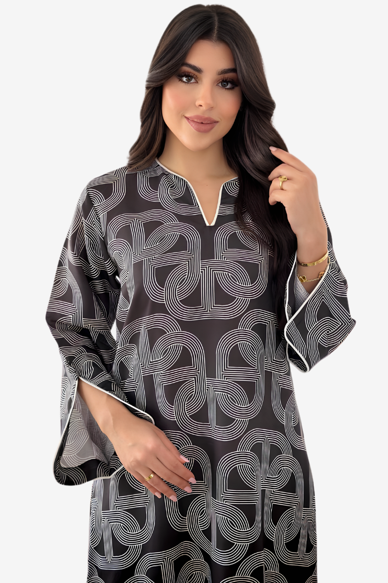 A woman modeling a black long-sleeve tunic with a white geometric interlocking pattern and white piping details on the neckline and cuffs, set against a plain light gray background.