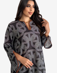 A woman modeling a loose-fitting black tunic with a white geometric interlocking line pattern and a split neckline, photographed against a light gray studio background.