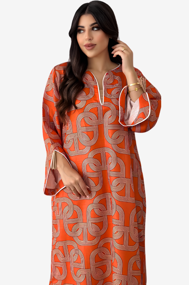 A woman modeling a long-sleeved orange kaftan dress with a beige geometric interlocking pattern, featuring a split neckline with white drawstrings and flared bell sleeves with white piping.