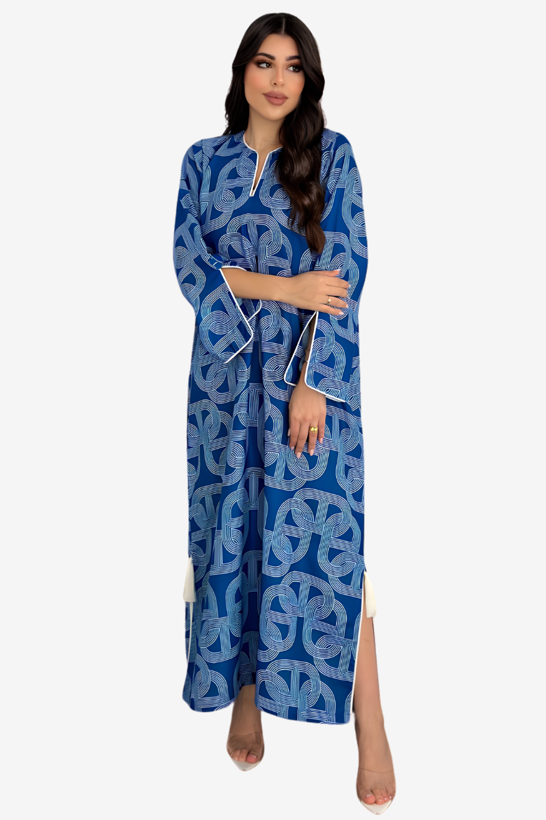 A woman modeling a long blue kaftan dress with a white geometric pattern and white piping details, set against a plain light gray background.