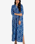 A woman modeling a long blue kaftan dress with a white geometric pattern and white piping details, set against a plain light gray background.