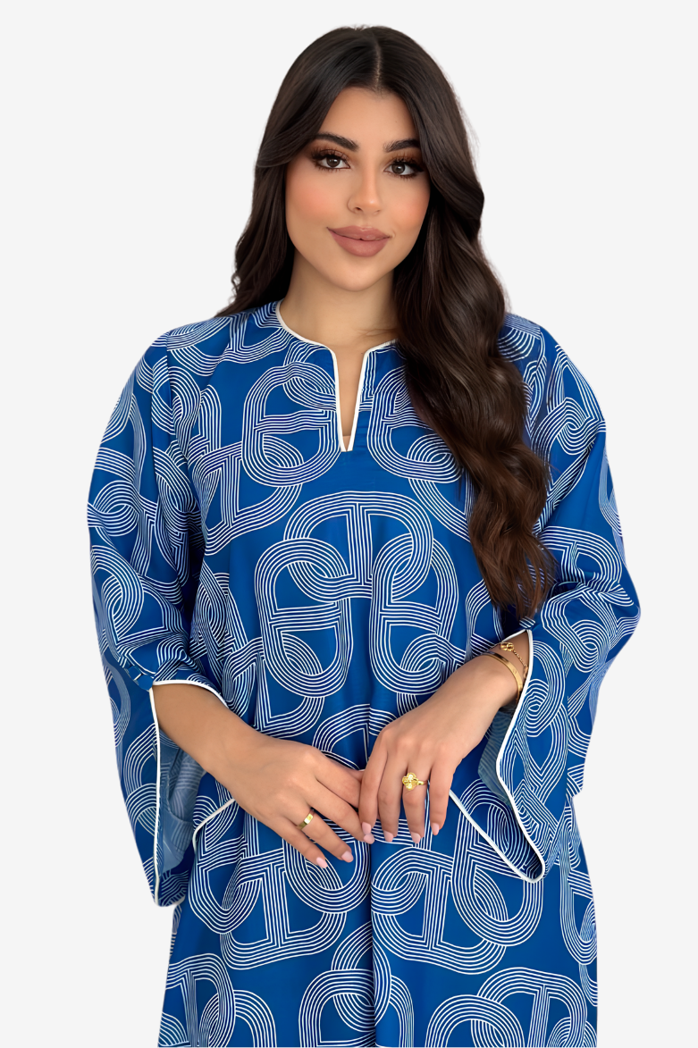A woman modeling a blue kaftan dress with a white geometric pattern and white piping details on the neckline and cuffs, set against a plain light gray background.