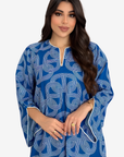 A woman modeling a blue kaftan dress with a white geometric pattern and white piping details on the neckline and cuffs, set against a plain light gray background.