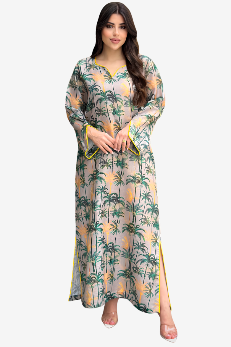 A woman modeling a long-sleeved maxi dress with a tropical palm tree print on a light beige background. The dress features green and yellow palm trees, yellow sun motifs, and yellow piping details along the cuffs and side slits, paired with clear high-heeled sandals.