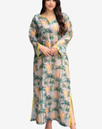 A woman modeling a long-sleeved maxi dress with a tropical palm tree print on a light beige background. The dress features green and yellow palm trees, yellow sun motifs, and yellow piping details along the cuffs and side slits, paired with clear high-heeled sandals.