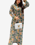 A woman modeling a long-sleeved maxi dress with a green palm tree print on a beige background, holding a white handbag with a gold LV logo.