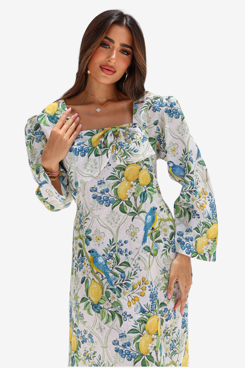 A woman modeling a long-sleeve midi dress with a white base and a vibrant lemon, blue bird, and floral print, set against a plain light gray background.