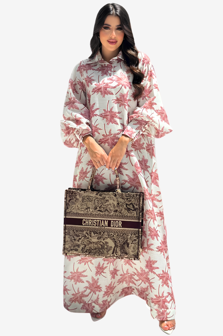 A woman modeling a long-sleeved, high-neck maxi dress with a pink palm tree pattern on a white background, holding a Christian Dior tote bag.