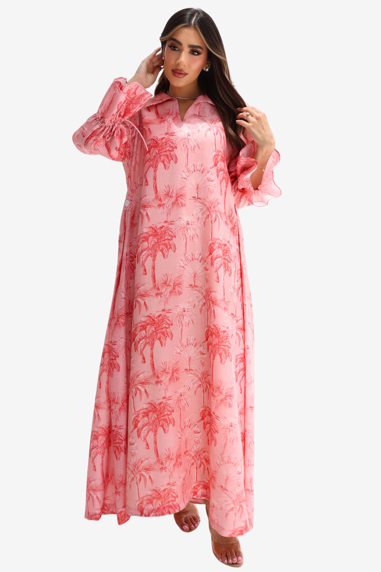 A woman modeling a long pink maxi dress with a red palm tree print, featuring ruffled sleeves and a collared neckline, set against a plain light gray background.