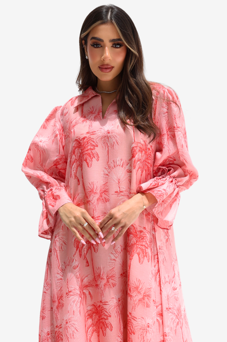 A woman modeling a pink long-sleeve shirt dress with a red palm tree pattern, featuring a collar and voluminous bell sleeves, set against a plain light gray background.