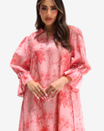 A woman modeling a pink long-sleeve shirt dress with a red palm tree pattern, featuring a collar and voluminous bell sleeves, set against a plain light gray background.