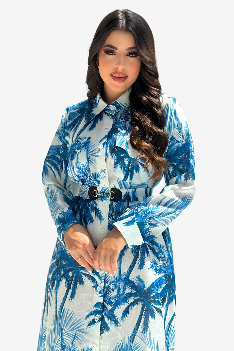 A woman modeling a long-sleeve collared shirt dress with a blue and white palm tree print, featuring a matching fabric belt with a black buckle, set against a plain light gray background.