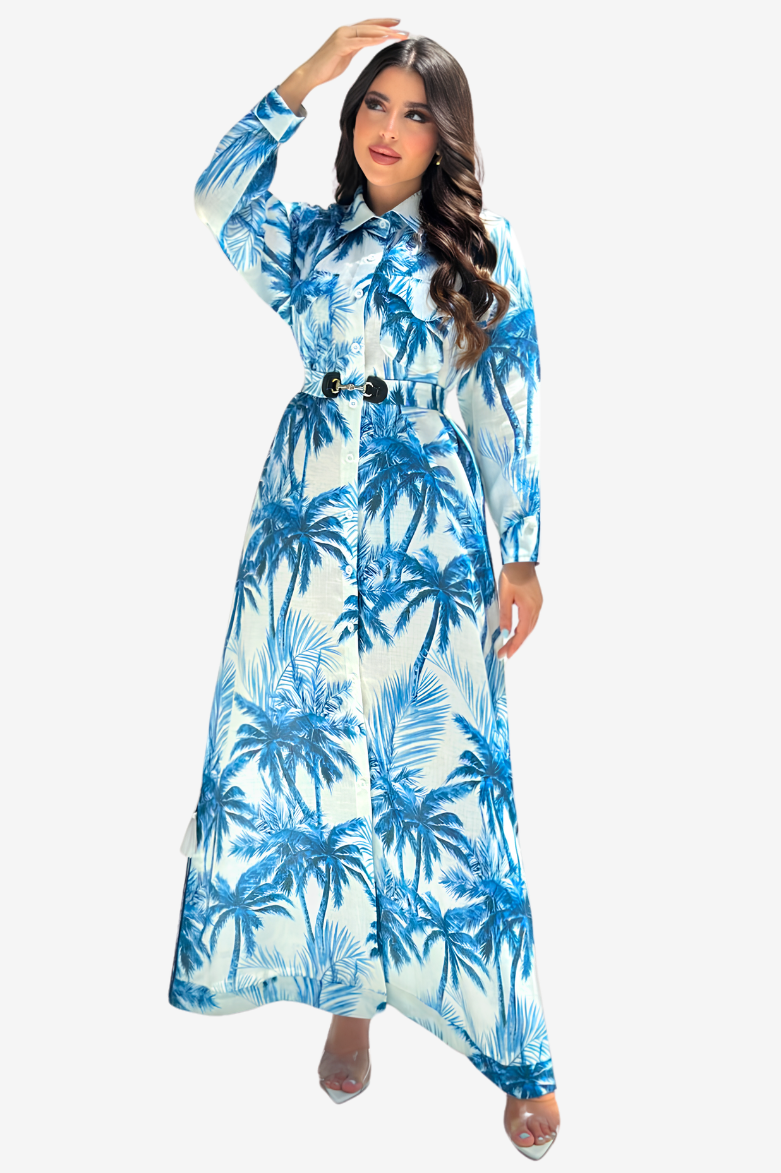 A woman modeling a long-sleeve, collared maxi dress with a blue and white palm tree print, featuring a matching belt with double gold-tone buckles, set against a plain light gray background.