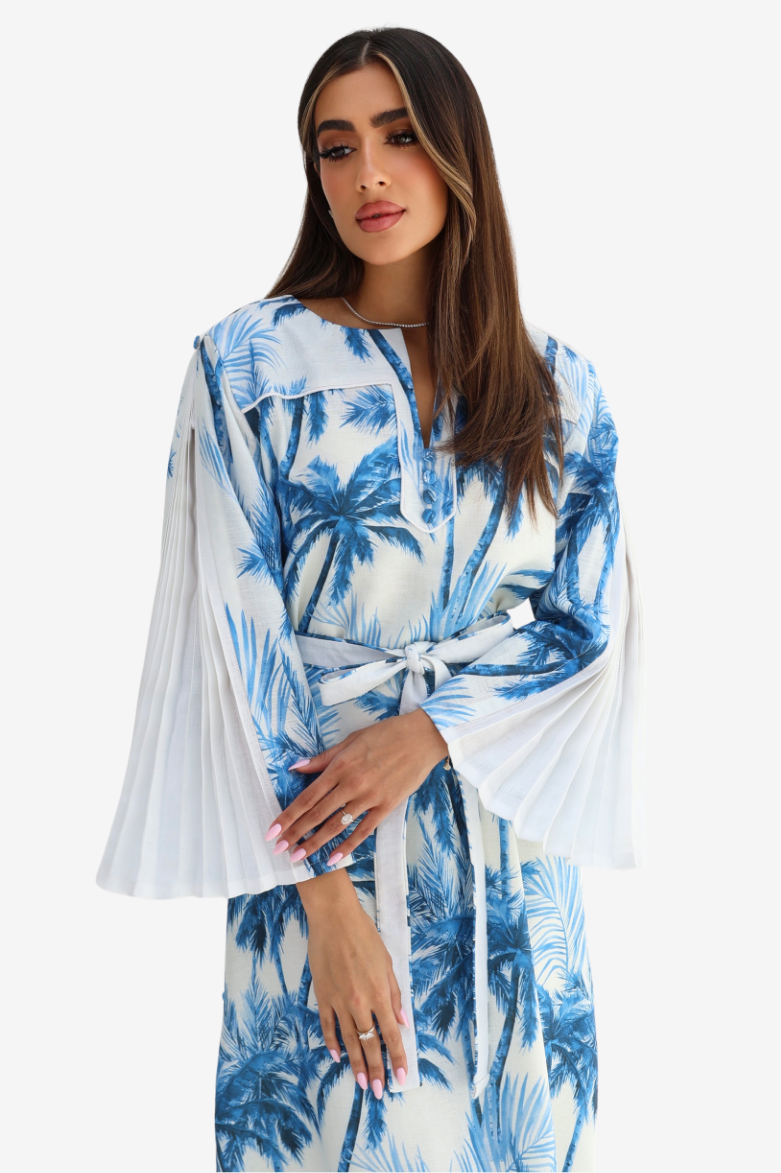 A woman wearing a white maxi dress with a blue palm tree print, featuring wide flared bell sleeves and a matching waist tie, set against a plain light gray background.