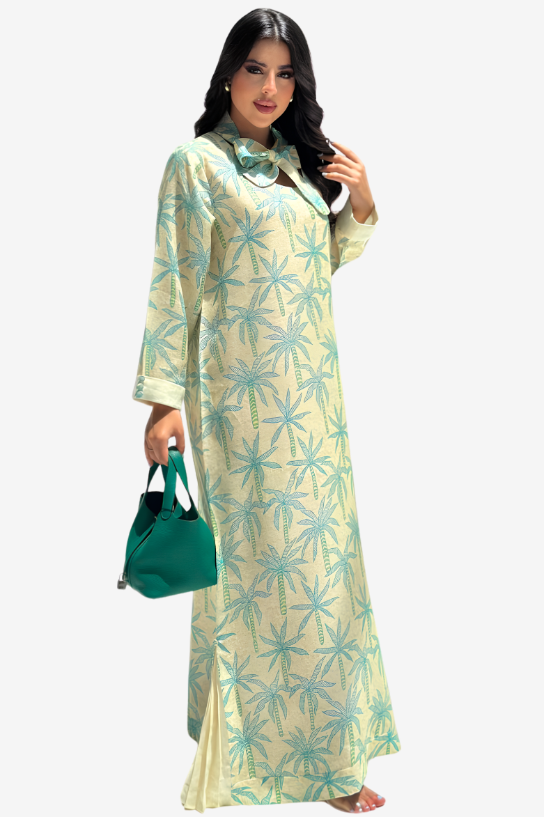A woman modeling a long-sleeved, floor-length cream-colored dress with a light blue palm tree pattern, holding a green handbag against a plain light gray background.