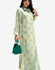 A woman modeling a long-sleeved, floor-length cream-colored dress with a light blue palm tree pattern, holding a green handbag against a plain light gray background.